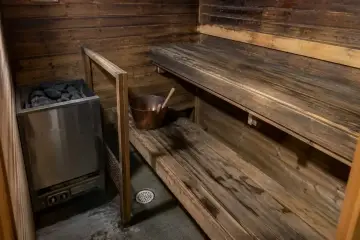 Stará dřevěná sauna před rekonstrukcí s tmavým opotřebovaným obložením, zašlými lavicemi, elektrickými kamny s kameny a vědrem s naběračkou.