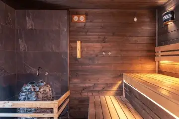 Interiér sauny s dřevěnými lavicemi, kamennou stěnou a saunovými kamny.